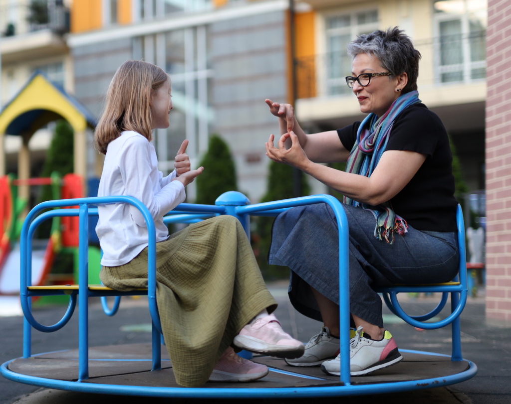 A young girl and an older woman joyfully mirror hand gestures, resembling sign language, while seated on a blue merry-go-round at an outdoor playground.